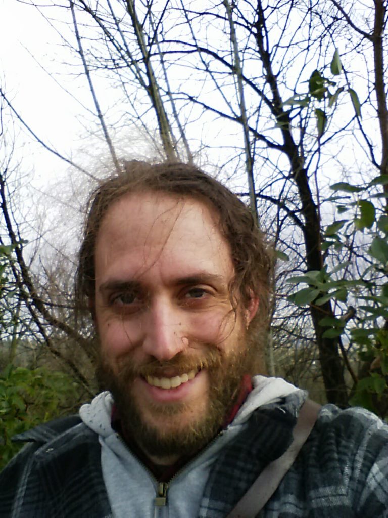 An image of the author smiling, with bare trees and a few leafy shrubs in the background. Wind is tugging his hair into fly-aways, and it is raining around him despite the bright grey sky.