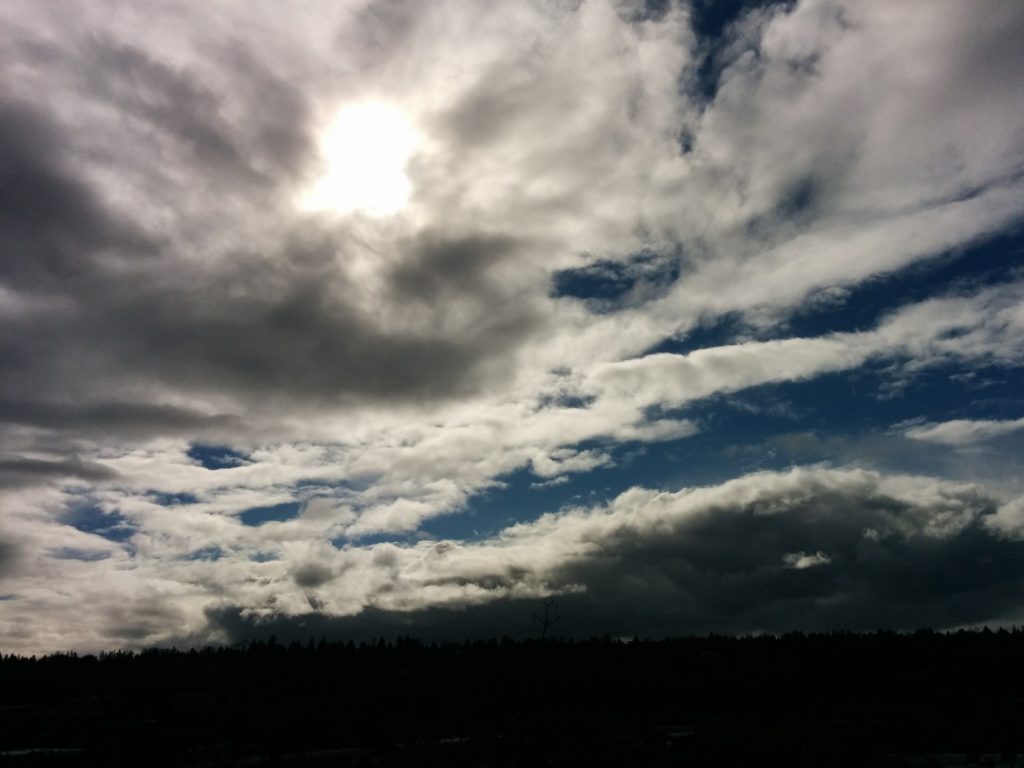 A chaotic bunch of clouds stream across the sky, the sun shining through a large clump of them.