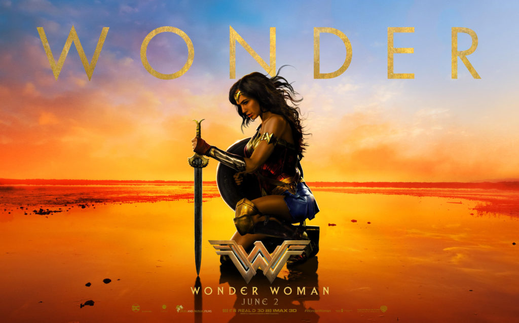 An image of Gal Gadot as wonder woman, kneeling on a beach at sunset. Her sword is planted in the ground in front of her, her left hand on the hilt. She holds her shield in her right hand. She is wearing her full armor, and her hair is teased lightly in the breeze. Her head is slightly bowed, as if in contemplation.