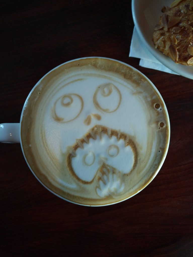 A cup of coffee with latte art depicting, as far as I can tell, a head eating another head with its fangs.
