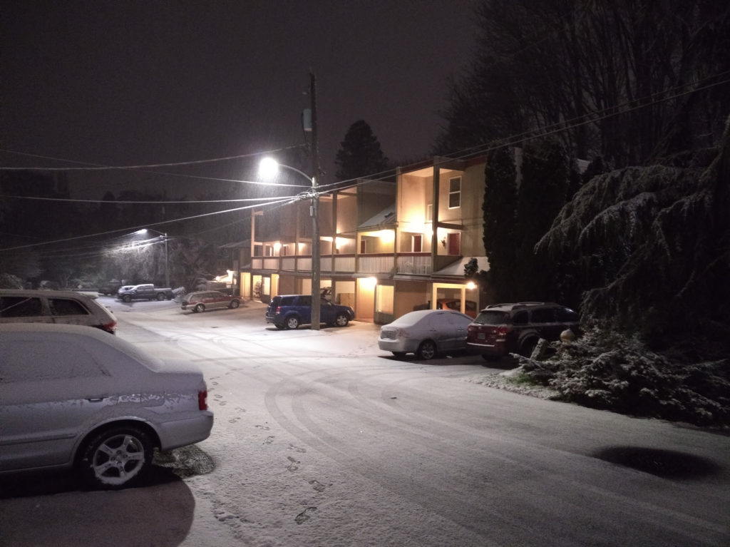 An image of a snow-covered street, with an apartment building in the background, lit by streetlights.