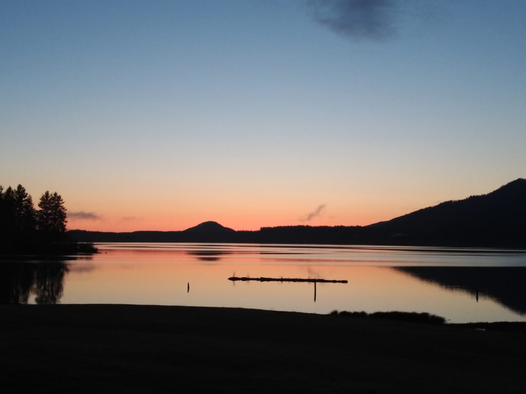 A picture of a sunset, just before dark, as the sun descends below the horizon. The colors reflect off a lake that takes up most of the foreground, though you can see the shore as well.