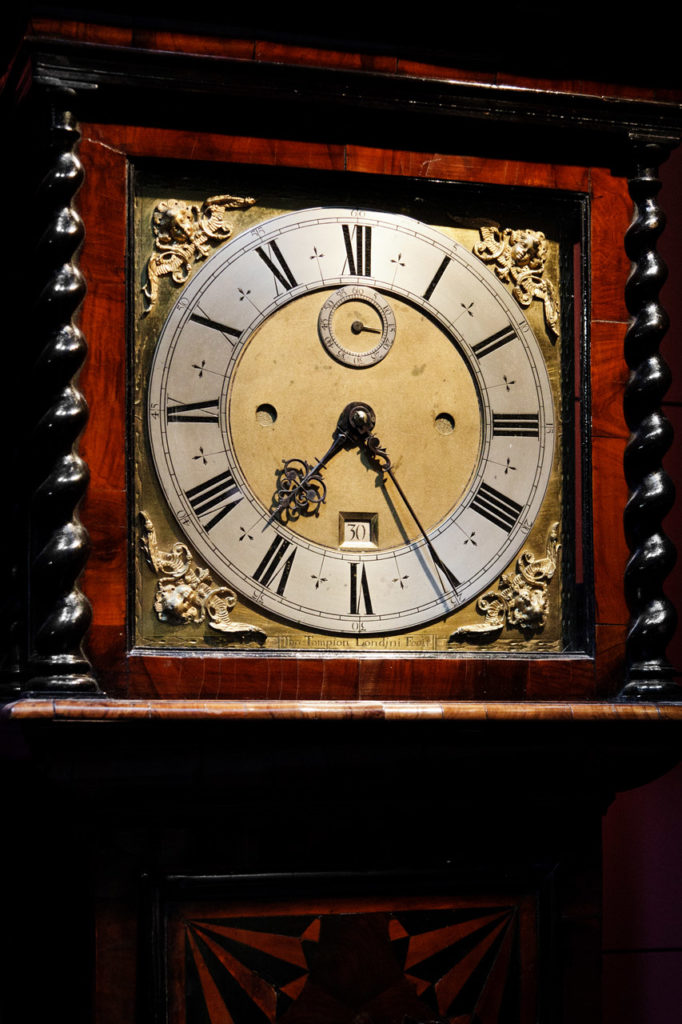 An antique clock, with wood frame, brass fittings, and a silver circle containing the numbers. It currently reads 7:25, minute hand pointed at the five, hour hand just past the seven.