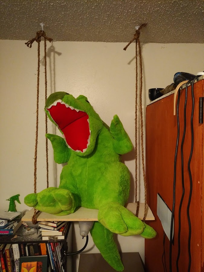 A three foot tall plush dinosaur sits in a handmade wood-and-rope swing, suspended from a bedroom ceiling, with its back to the wall and its face facing the camera. It has a happy, wide-open mouth.