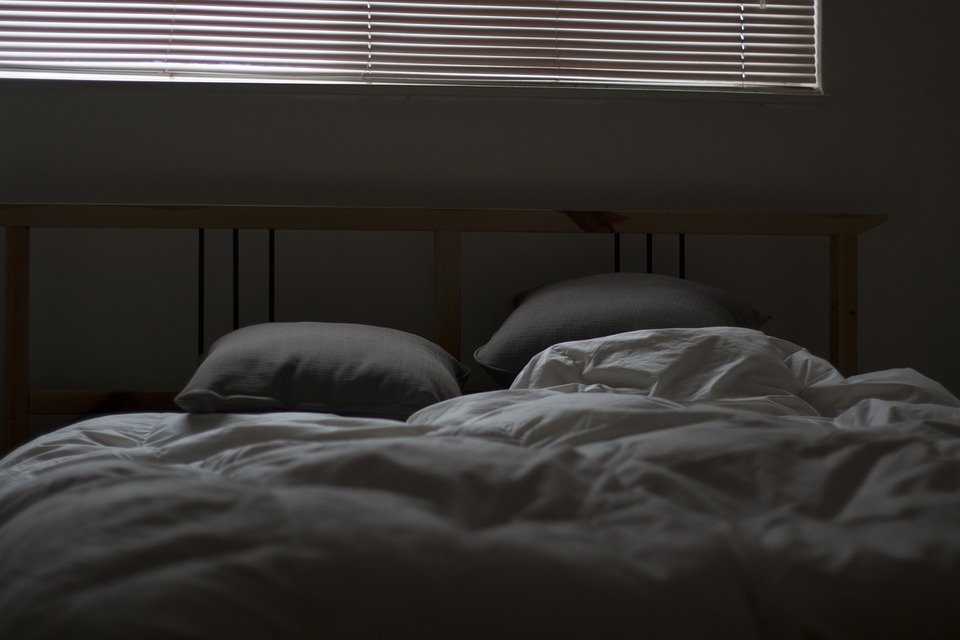 A dimly lit bedroom. In the top of the frame, light filters though the blinds. It illuminates a bed, covered in rumpled white sheets and a couple pillows.