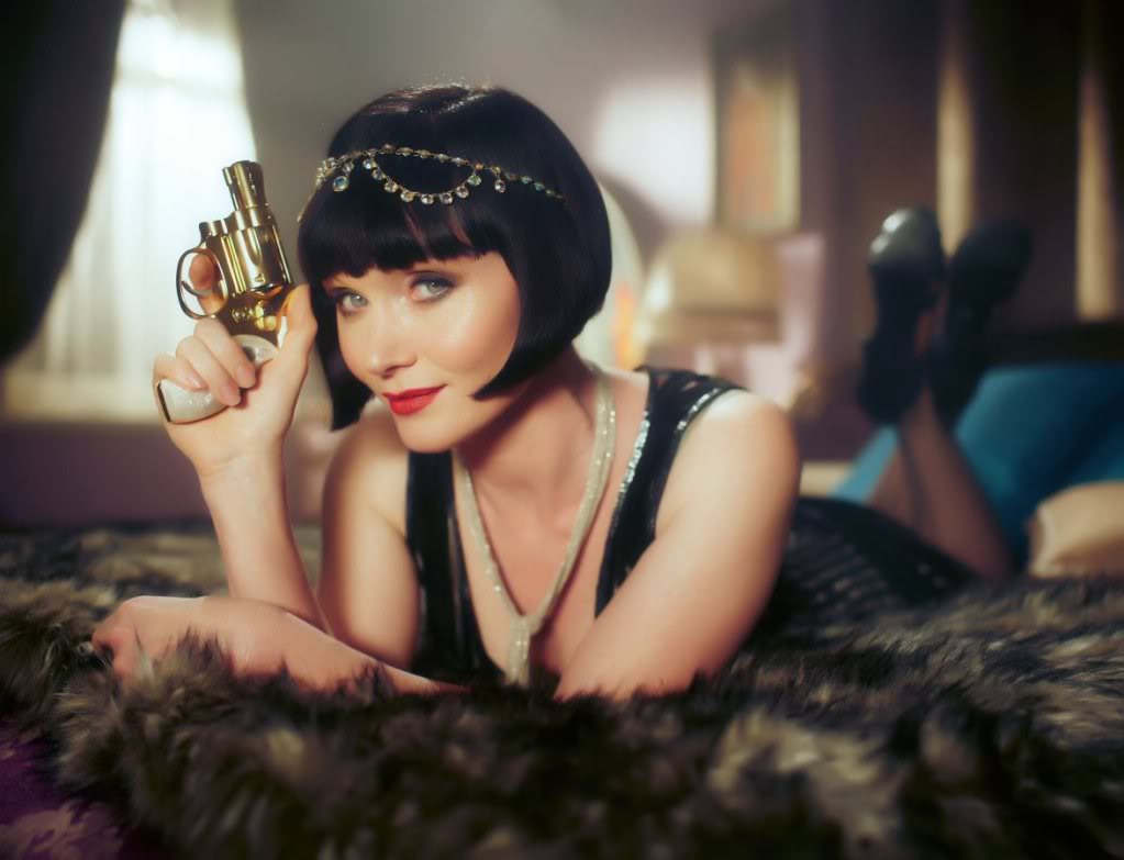 An image of Phryne Fisher, the rich dilettante flapper protagonist of Miss Fisher's Murder Mysteries, laying on a bed, holding up a gold snub-nosed revolver, and looking coquettishly at the camera.