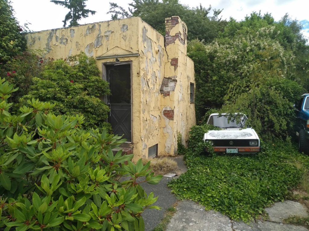 A building with crumbling yellow paint that flakes off the brickwork. A beat-up old volkswagon is parked behind it, half-covered by the bushes that have grown over, under, and around it.