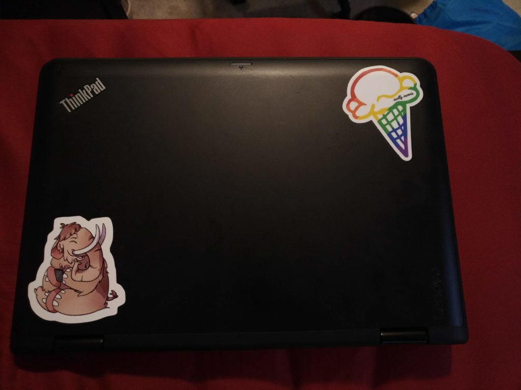 Image of a closed 11" thinkpad's lid with a Mastodon sticker stuck in the lower-left corner and a rainbow ice cream cone sticker from Molly Moon's in the upper right.