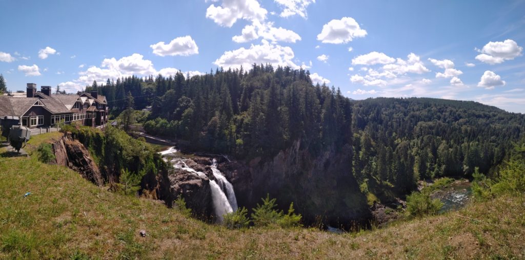A panoramic shot of Snoqualmie Falls in the state of Washington. Twin waterfalls fall over a high cliff and merge together before crashing into the river below. The rest of the valley is lined with trees.