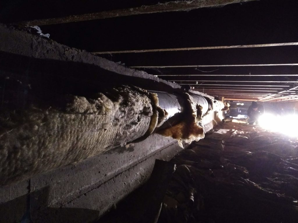 Photo taken in the crawlspace under a house. The area is dimly lit by spotlights, illuminating the dirt floor and a vent covered with ragged insulation. The joists overhead are cloaked in shadow.