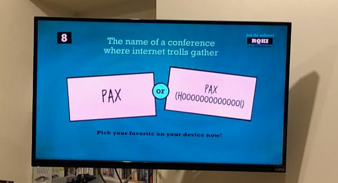A game of Quiplash 2 on Jackbox Party Pack 4, displayed on a hanging flatscreen TV. The prompt reads "The name of a conference where internet trolls gather." The answers provided by the players are "PAX" and "PAX (HOOOOOOOOOOOOO!)."