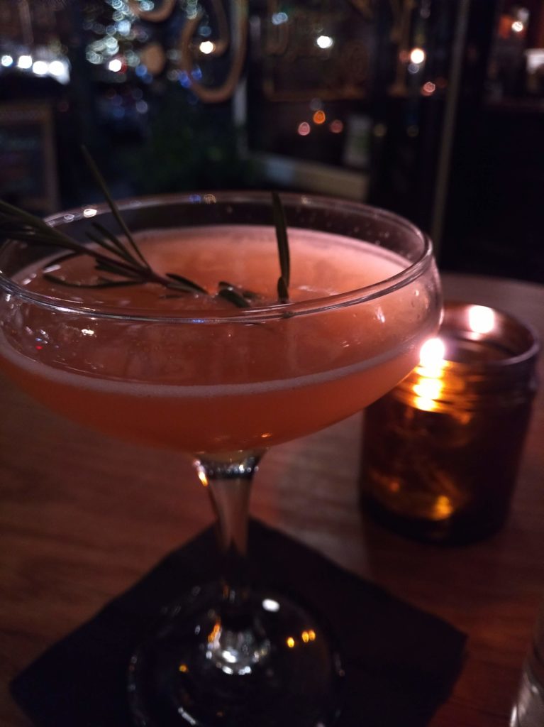 A orange/pink cocktail with a sprig of rosemary in it sitting in front of a candle on a table.