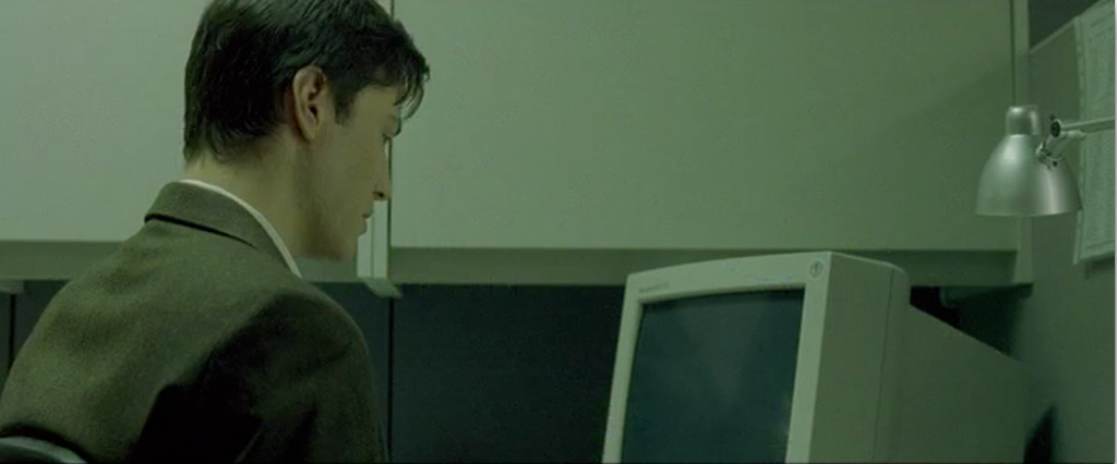 Neo from the Matrix, in an ill-fitting suit, looking at a monitor in his work cubicle.