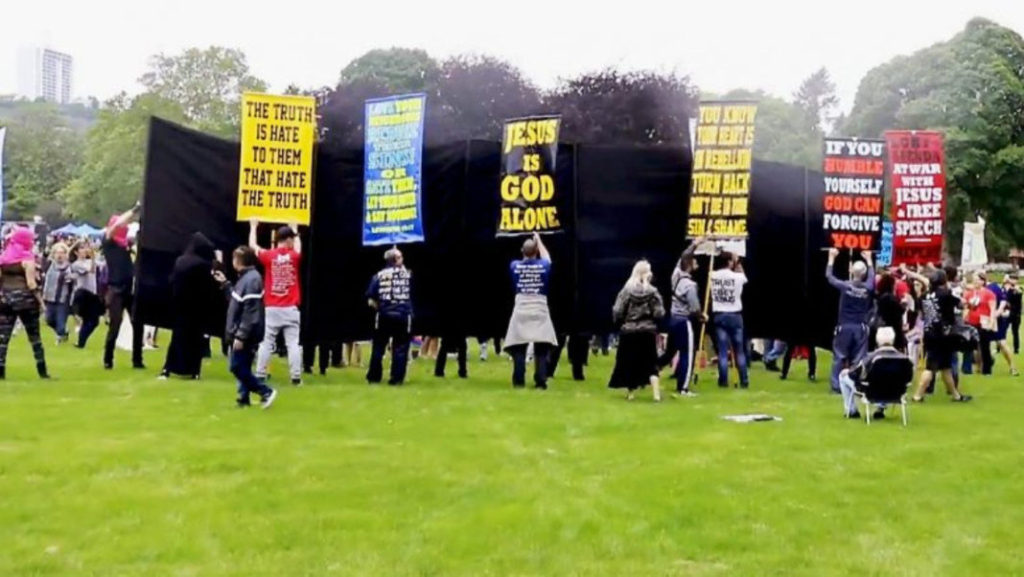 Alt-right protestors holding up signs. In front of them is a large black wall-like structure being held up by pink t-shirt mask wearing protestors.