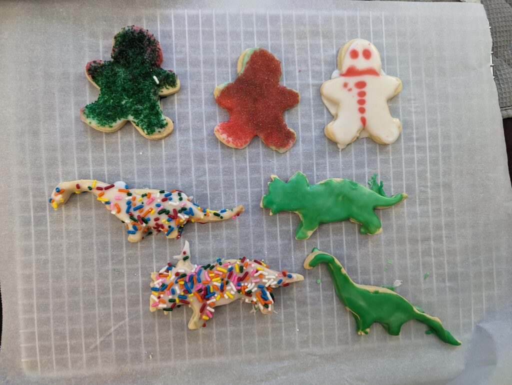 Homemade Christmas cookies in the shape of gingerbread men and dinosaurs on parchment paper on a cooling tray. The icing and sprinkles are a bit messy.