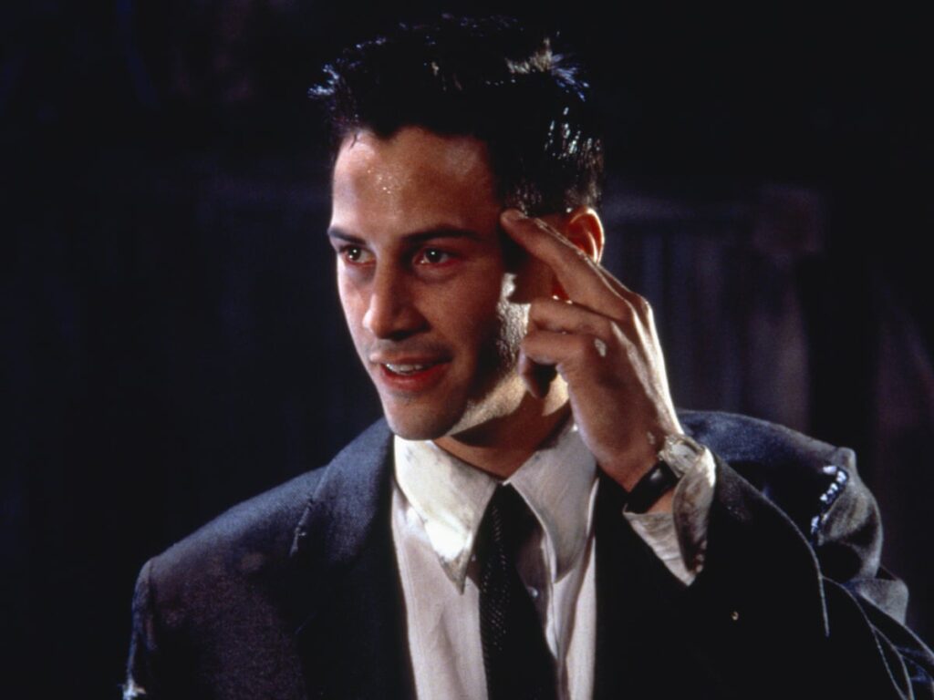 A still of Keanu Reeves from Johnny Mnemonic. He's tapping the side of his head with two fingers, and looks roughed up and a little crazed.