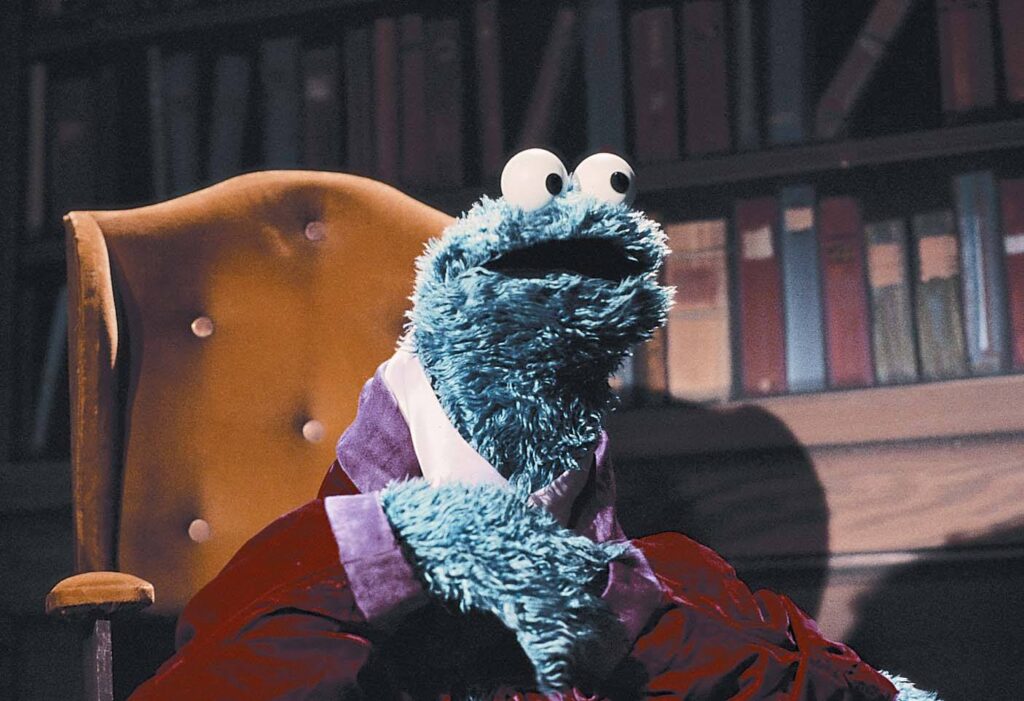 An image of Cookie Monster sitting in a chair in front of shelves of books from the Monsterpiece Theater segment for Sesame Street.