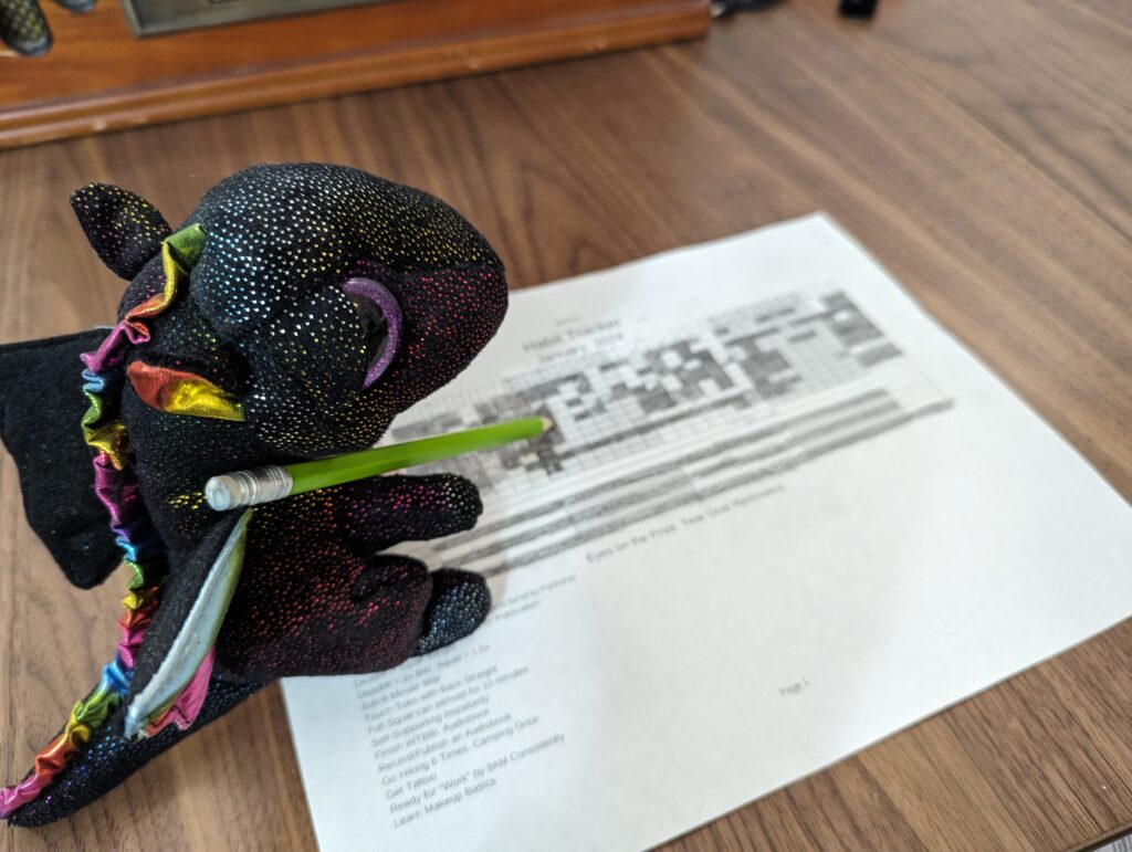 A stuffed dragon holding a pencil, writing on a habit tracker.