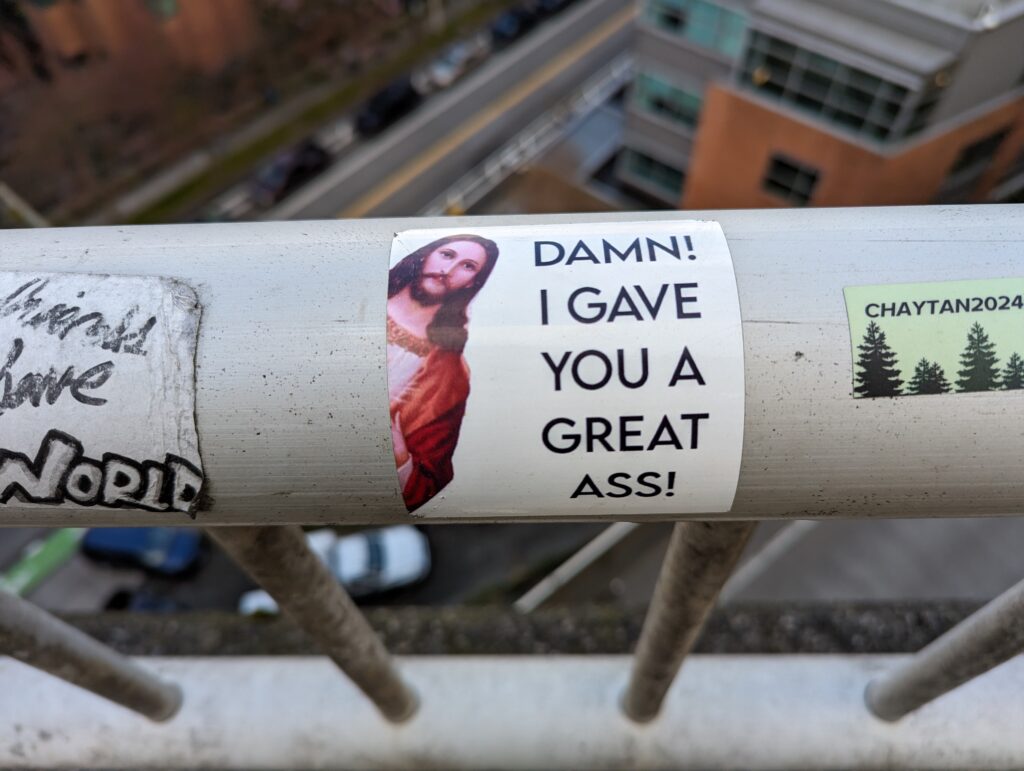 A sticker attached to a bridge handrail. Jesus is leaning in from the left side of the sticker and the text reads "Damn! I gave you a great ass!"