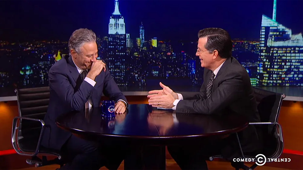 An image of Stephen Colbert interviewing Jon Stewart on The Colbert Report. Colbert is grinning and Stewart is laughing into his hand.